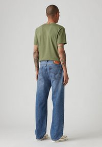 Άνδρας με τατουάζ φορώντας ελιά πράσινο φανελάκι, χαλαρούς μπλε τζιν και λευκά αθλητικά παπούτσια, ο οποίος είναι γυρισμένος προς τα πίσω σε ουδέτερο φόντο.