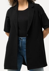 Svart kortärmad kavaj med slagkrage, enkelknäppt stängning och sidofickor, bärs över en svart topp och blå jeans.