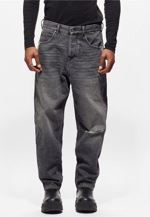 Mann trägt ein schwarzes langärmliges Oberteil, locker sitzende, ausgewaschene schwarze Jeans mit Knie-Riss und schwarze klobige Stiefel, steht vor einem einfarbigen Hintergrund.