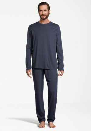 Haut de pyjama à manches longues en bleu marine et pantalon assorti en tissu doux, avec un col rond et une coupe décontractée. Pieds nus sur un fond blanc.