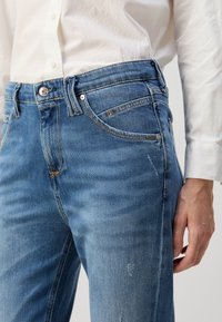 Světle modré džíny z denimu s mírným vyblednutím, pěticípým designem a kovovým knoflíkem. Tkanina má hladkou texturu s minimálním opotřebením.