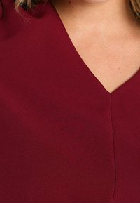 Femme portant un haut bordeaux à col en V, montrant la partie supérieure de la poitrine et des épaules avec une peau claire et des mèches de cheveux blonds.