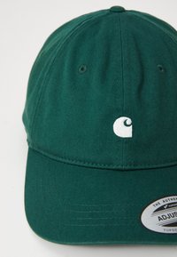 Green cotton baseball cap with a curved brim, featuring a white embroidered logo and an adjustable strap at the back, showing textured fabric.
