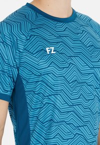 Blå kortärmad sportskjorta med mörkblå vinklad linjemönster och vitt "FZ"-logotyp på övre bröstet, bar av en person.