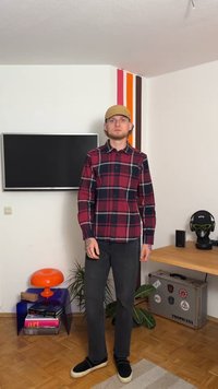 Raudona ir tamsiai mėlyna languota ilgomis rankovėmis marškiniai, juodos džinsai ir juodos šlepetės; ant rankų tatuiruotės; ant galvos paprasta beisbolo kepuraitė.