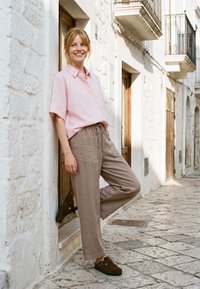 Femme en chemise rose et pantalon beige appuyée contre un mur de pierre blanche dans une rue étroite ensoleillée pavée de pierres.