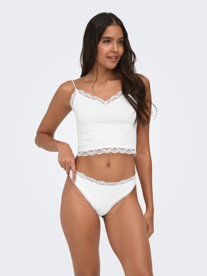 Młoda kobieta o długich ciemnych włosach, ubrana w biały top z koronkowymi wykończeniami i pasujące majtki, pozująca z ręką na biodrze.