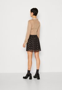 Camisa longa transparente em tom bege com gola, combinada com uma saia preta com padrão de bolinhas. Sapatos pretos de salto grosso completam o visual.
