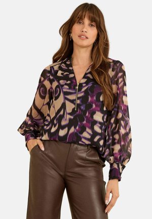 Blusa viola e nera con motivo, con zip frontale, maniche lunghe a sbuffo e una texture leggera e fluida, abbinata a pantaloni di pelle marroni.