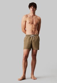 Bañador caqui con cintura elástica y cordón, con una textura suave y un bolsillo lateral. El modelo está descalzo frente a un fondo gris.