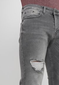 Jeans de mezclilla gris con un diseño desgastado y un gran parche rasgado en la rodilla. La tela tiene una textura suave con un desvanecimiento sutil.