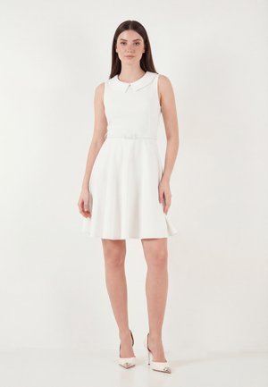 Žena stojí, oblečená v bielej šatke bez rukávov s golierom Peter Pan, opásaná v páse a v bielych vysokých podpätkoch na jednoliatom bielom pozadí.