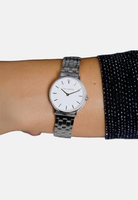 Wrist wearing a silver metal bracelet watch with a white face, black hour markers, and "Paul Valentine" text, time showing 10:10.
