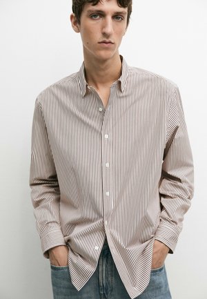 Jonge man die een beige en bruine smal gestreepte blouse met knopen en lange mouwen en een blauwe spijkerbroek draagt, staand met handen in de zakken.