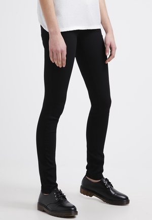 Personne portant un pantalon noir slim, des chaussures noires à lacets avec des semelles épaisses, et un haut blanc texturé à manches courtes, debout sur un sol blanc.