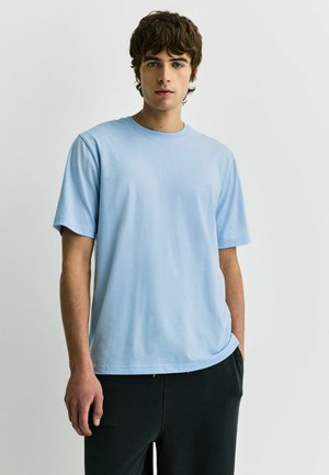 Ung mand med kort krøllet hår iført en lyseblå t-shirt og sorte bukser, stående mod en ensfarvet hvid baggrund.