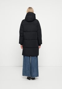 Manteau matelassé noir avec capuche, à la texture matelassée et de longueur longue, associé à un pantalon en denim à jambes larges.