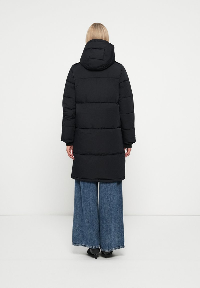 Manteau matelassé noir avec capuche, à la texture matelassée et de longueur longue, associé à un pantalon en denim à jambes larges.