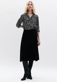 Zwarte wikkelrok en zwart-wit gestippeld blouse met opgerolde mouwen. Model draagt knielange zwarte laarzen en een gouden kettinghalslace.