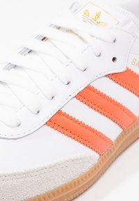 Vitt sneakers med orange ränder, läderöverdel, texturerad mockatå och kontrasterande sula i gumgummi. Har sydda detaljer och snörning.