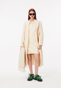 Cappotto oversize crema, vestito a maglia fitted crema con costine, occhiali da sole neri e sneakers vivaci verdi e nere con un dettaglio di lacci unico.
