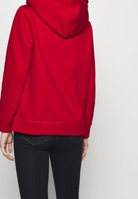 Červená mikina s kapucí se žebrovanými manžetami a lemem, má volný střih a měkkou texturu. Tmavě modré džínové kalhoty s částečně viditelnými zadními kapsami.