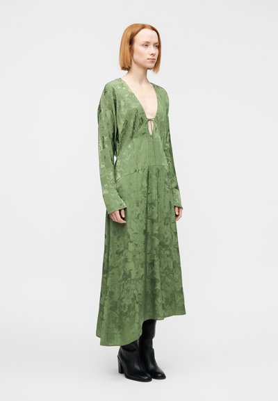 Πράσινο maxi φόρεμα με υφή σχεδίου, βαθύ V-λαιμόκοψη, μακριά μανίκια και κλεί σιμο με φιόγκο στο στήθος, συνδυασμένο με μαύρες μπότες με τακούνι.