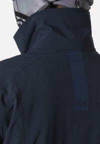 Námořnická modrá voděodolná bunda s vysokým límcem, matným povrchem, štíhlým střihem a logem značky z kontrastního materiálu.