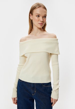 Cremefarbener, gerippter Off-Shoulder-Pullover mit langen Ärmeln, der über einen breiten, umgeschlagenen Ausschnitt und einen taillierten Oberkörper verfügt, kombiniert mit dunkelblauen Jeans.
