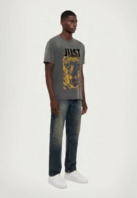 Grå T-skjorte med gul tigergrafikk og svart tekst. Kombinert med falmede blå jeans og hvite joggesko. Modellen står vendt mot høyre.