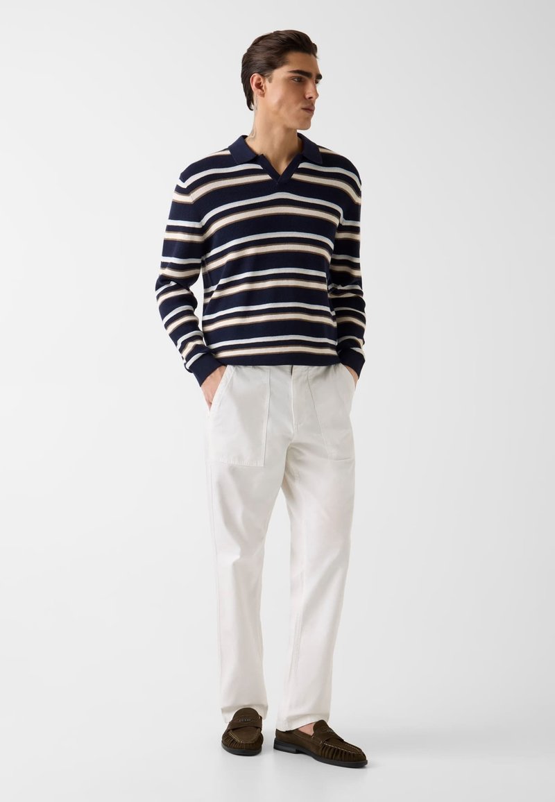 Homme debout les mains dans les poches portant un pull rayé bleu marine, un pantalon blanc et des mocassins marron sur un fond clair uni.