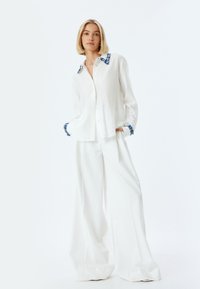 Camisa blanca con cuello y puños estampados en azul, combinada con pantalones blancos de pierna ancha. Textura de tela suave, ajuste relajado.