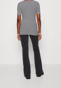 Persona vestida con una camiseta de manga corta de rayas en blanco y negro, pantalones negros y zapatos negros, de pie mirando hacia atrás contra un fondo sencillo.