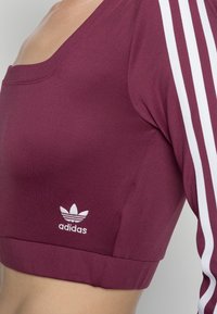 Vínové zkrácené tričko vyrobené z elastického materiálu. Má čtvercový výstřih, bílé tříproužkové provedení na rukávech a bílé logo Adidas na dolním lemu.