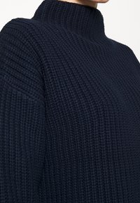 Granatowy dzianinowy sweter z wysokim, prążkowanym kołnierzem. Teksturowana tkanina ma grube rowki, które nadają mu strukturalny wygląd.