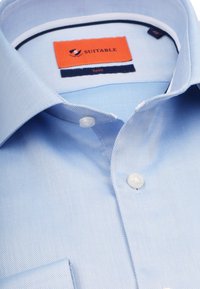 Lichtblauw overhemd van keperstof, voorzien van een standaardkraag, witte knopen en een label met de tekst "SUITABLE".