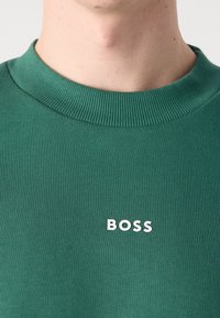 Zelený svetr s kulatým výstřihem a žebrovanými okraji. Na hrudi se nachází bílé logo "BOSS", vyrobený z hladkého a měkkého materiálu.