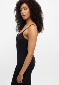 Μαύρο τοπ με λεπτές τιράντες, με υφή υφάσματος και εφαρμοστή σχεδίαση. Το μοντέλο εμφανίζεται από το πλάι, φορώντας χαλαρό μαύρο παντελόνι.