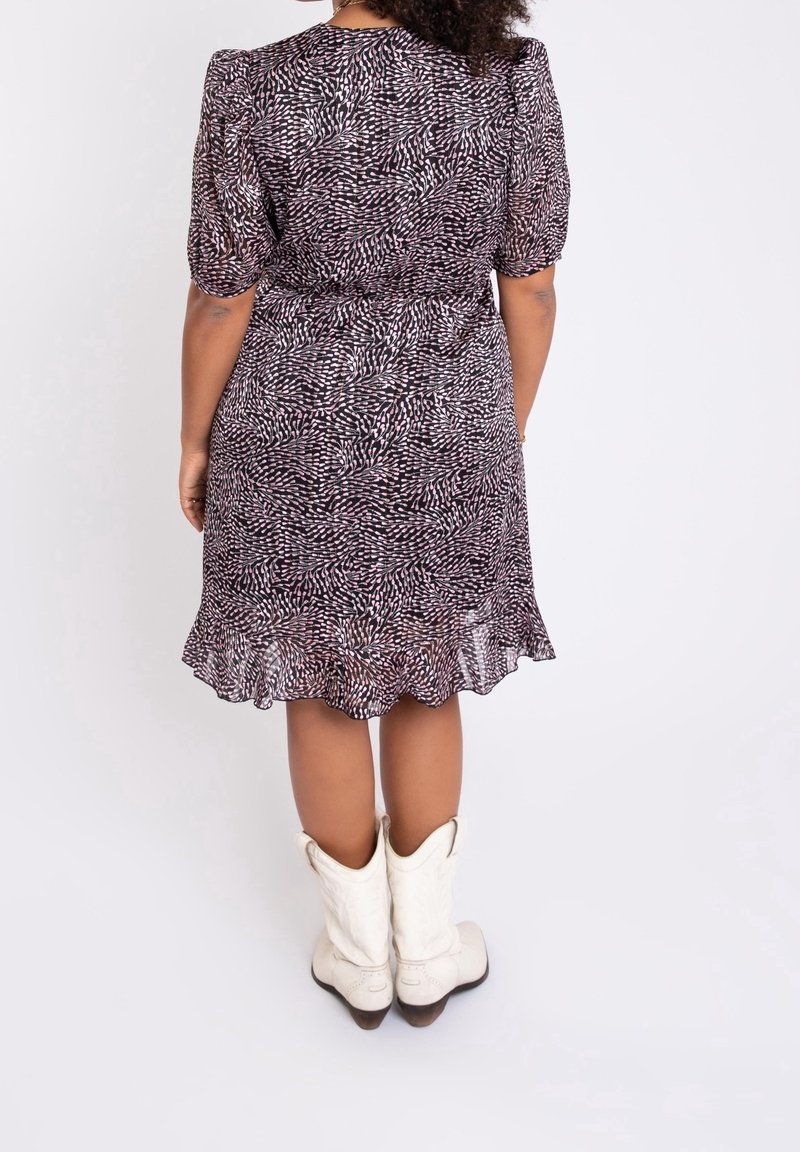 Femme portant une robe violette à motifs, longueur genou, manches courtes, et des bottes de cowboy blanches, debout de dos.