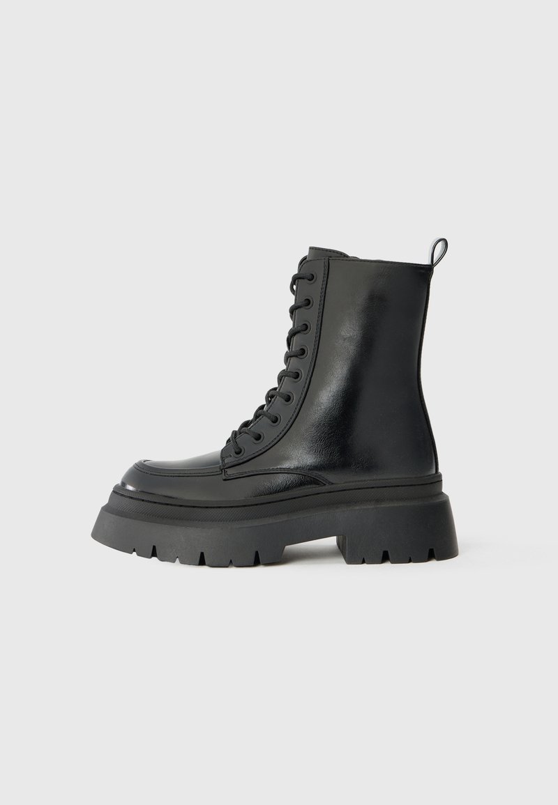 Botte de combat noire montante avec semelle épaisse à crampons, fermeture à lacets sur le devant et languette de préhension sur fond gris.
