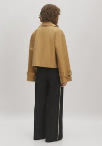 Ingekorte, tan trenchcoat met een brede kraag en gefronteerde mouwen. Gecombineerd met zwart, hooggesneden broek met een verticale witte streep.