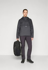 Zwarte geïsoleerde pulloverjas met een halve rits, gecombineerd met donkere broek en zwarte laarzen. Een zwarte rugzak wordt in één hand vastgehouden.