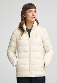 Beige pufferjacka med dragkedja framtill, hög krage och mönster av horisontella kanaler. Har sidofickor och en logotyp på bröstet.