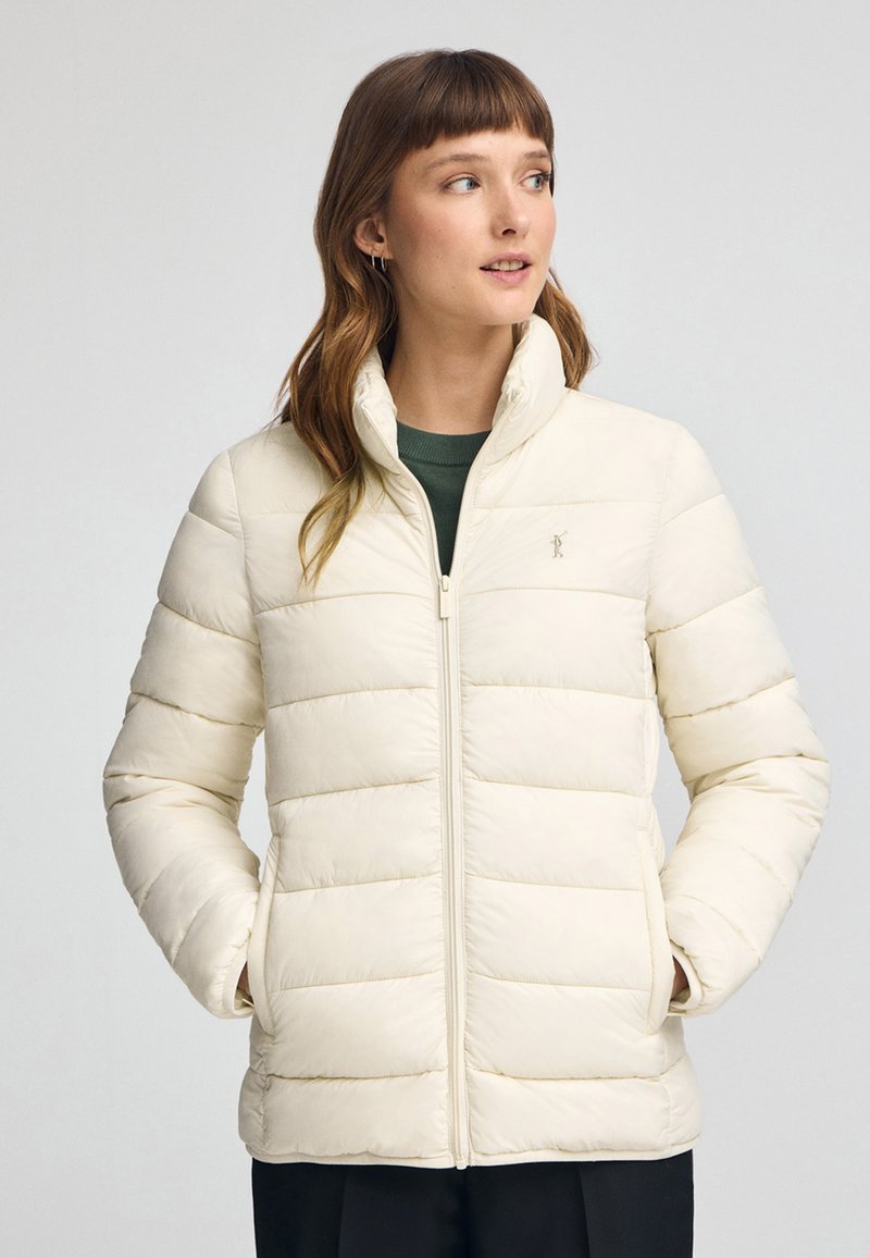 Beige pufferjacka med dragkedja framtill, hög krage och mönster av horisontella kanaler. Har sidofickor och en logotyp på bröstet.