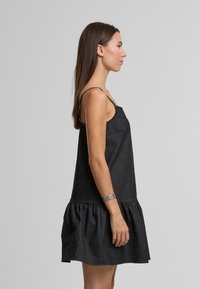 Melnas denima kleita ar plānām plecu siksnām, slāņainu volānu apakšmalu un gludu tekstūru. Sānu skatījumā redzama tās brīvā piegriezuma forma.