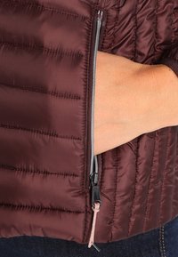 Bordeauxrode gewatteerde jas met een zijritszak. Glad, glanzend stof, geaccentueerd met een zilveren rits en een roze trekkoorddetail.