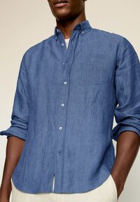 Blaues Leinenhemd mit Button-Down-Kragen, langen hochgekrempelten Ärmeln, einer einzelnen Brusttasche und einem abgerundeten Saum. Weiche Textur und leicht.