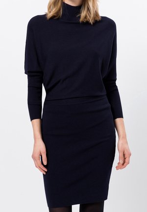 Kobieta w dopasowanej czarnej dzianinowej sukience z długim rękawem i wysokim kołnierzem na jednolitym białym tle.