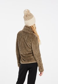 Giacca in pile marrone con colletto alto, abbinata a un berretto in maglia beige con pompon in cima. Pantaloni non visibili.