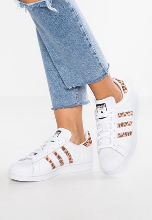 Personne portant un jean bleu court effiloché et des baskets blanches Adidas avec des bandes et un détail au talon imprimés léopard sur fond blanc.